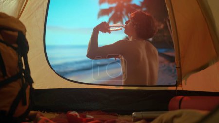 Téléchargez les photos : Le concept de camping et d'aventure. Un homme est en vacances de camping relaxant au bord de l'océan. Jeune homme assis près d'une tente au coucher du soleil sur la plage boire de l'eau d'une bouteille. Vacances dans la nature. - en image libre de droit
