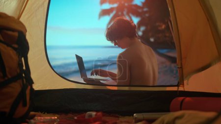 Téléchargez les photos : Concept de Camping et Aventure. Un homme est en vacances de camping relaxant au bord de l'océan. Jeune homme assis près d'une tente au coucher du soleil sur la plage travaillant sur un ordinateur portable. Vacances dans la nature. - en image libre de droit