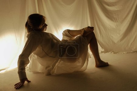 The silhouette of a dancer dancing on the floor against a light background is captured in motion by flowing fabrics.