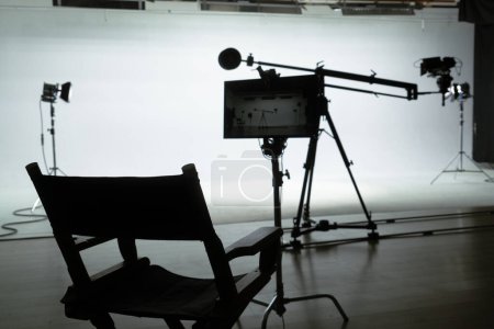 Photo for A directors chair silhouetted against the backdrop of a professional film set with lights and camera equipment. Backstage shot. - Royalty Free Image