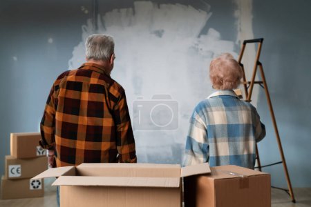 Téléchargez les photos : Un couple de personnes âgées évalue le travail inachevé dans leur maison, entouré de fournitures de peinture. Vue arrière. - en image libre de droit