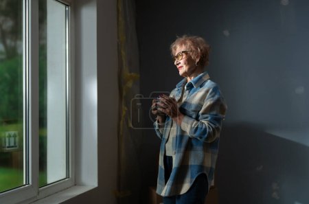 Téléchargez les photos : Femme âgée profite d'un moment de réflexion par la fenêtre au milieu des travaux de rénovation. - en image libre de droit