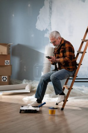Foto de Un hombre mayor se sienta en una escalera, profundamente absorto en su teléfono en medio de herramientas de pintura durante las renovaciones del hogar. - Imagen libre de derechos