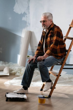 Photo for An elderly man sits on a ladder, immersed in his thoughts, holding a paint roller in his hands, among painting tools during a home renovation. - Royalty Free Image