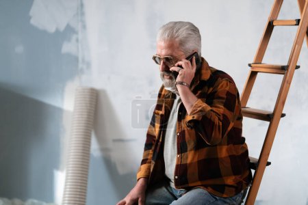 Photo for A senior man sits on a ladder, deeply engrossed in his phone amidst painting tools during home renovations. - Royalty Free Image