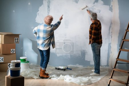 Téléchargez les photos : Une femme âgée supervise le processus de peinture des murs et montre à son mari comment faire le travail correctement. Vue arrière. - en image libre de droit