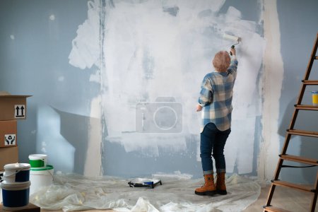 Photo for A focused elderly woman is immersed in the process of painting the walls of her home. Back view. - Royalty Free Image