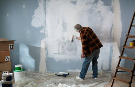 Téléchargez les photos : Un vieil homme concentré est plongé dans le processus de peinture des murs de sa maison. Vue arrière. - en image libre de droit