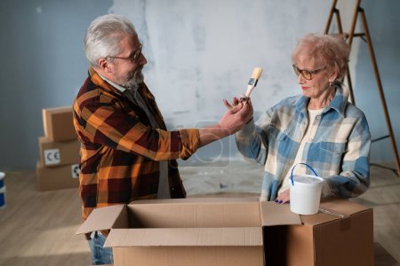 Foto de Una pareja mayor disfruta desempacando cajas durante una renovación del hogar, compartiendo un momento de alegría. Un anciano saca un pincel y felizmente se lo muestra a su esposa. - Imagen libre de derechos