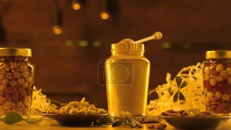 Foto de Miel orgánica saludable con nueces. Frascos de vidrio de pie sobre la mesa de madera con miel dorada fresca, cazo de miel colocada en el frasco abierto en el jarabe grueso, fondo de luz solar caliente. - Imagen libre de derechos