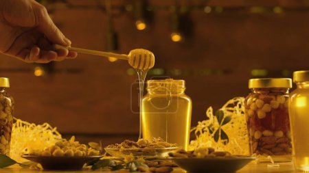 Photo for Healthy organic honey with nuts. Glass jars standing on the wooden table with fresh golden honey, man taking honey dipper and pours thick syrup over nuts, warm sunlight background. - Royalty Free Image