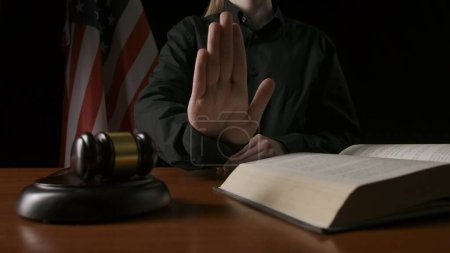 Foto de Ley y justicia. Mujer sentada en el escritorio con martillo de madera y libro de leyes, bandera de EE.UU. en el fondo. Juez mujer en la sala de la corte, muestra gesto de mano parada en la cámara. - Imagen libre de derechos