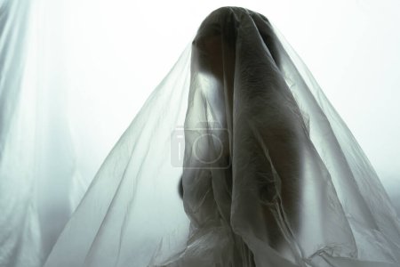 Photo for A ghostly silhouette of a figure stands covered by a sheer fabric, with a luminous white background creating an ethereal and mysterious effect. - Royalty Free Image