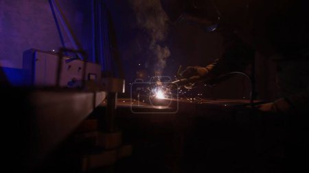 Foto de El proceso de un soldador que trabaja en un producto metálico. Soldador con máquina de soldadura en la superficie de hierro, humo, chispas y luces intermitentes alrededor. Ambiente oscuro con luz azul. - Imagen libre de derechos