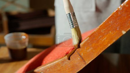 Großaufnahme einer Frauenhand, die mit einem Pinsel Flecken auf das Bein eines alten Holzstuhls aufträgt. Demonstration der Holzbearbeitung oder Möbelrestaurierung in einer Werkstatt.