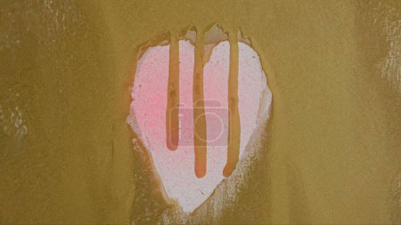 Téléchargez les photos : Une forme de coeur est révélée à travers une surface de verre peint en or, exposant un fond rose. Les couleurs et les textures contrastées créent un design visuellement attrayant et émotif. - en image libre de droit