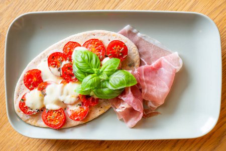 Foto de Mozzarella pita de tomate sobre pan plano con jamón fresco de Parma y decorado con hojas de albahaca servidas en un plato rectangular - Imagen libre de derechos