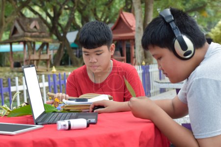 Photo for Young Asian boys doing homework, searching for information and do the reports on various species of tropical forests by using laptops, magnifiers, notebooks, pocket binoculars and tablets, soft focus. - Royalty Free Image