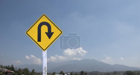 Señal de tráfico: Signo de giro en U izquierdo en poste de cemento junto a la carretera rural con fondo azul nublado blanco, espacio para copiar.