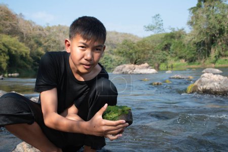 Photo for Asian schoolboy holding freshwater algae from diving into the river and pulling it up to study the fertility of the river's nature and including to do freshwater algae in his environment project work. - Royalty Free Image