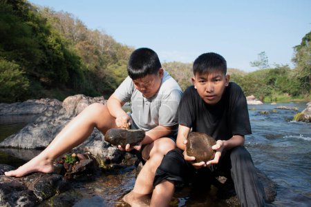 Photo for Asian boys're spending their freetimes by sitting on stones, holding rocks, scraping the top surface of stones to learn the natural life during their summer vacation in local national park. - Royalty Free Image