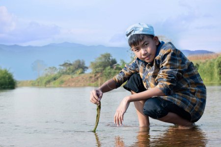 Photo for Asian boy in a plaid shirt wearing a cap holds freshwater algae that he plucks from a river in the area of his village to study the river's cleanliness, ecosystem, ecoeffect and pesticide effect. - Royalty Free Image