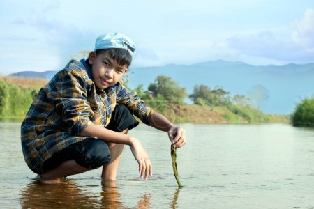 Téléchargez les photos : Asian boy in a plaid shirt wearing a cap holds freshwater algae that he plucks from a river in the area of his village to study the river's cleanliness, ecosystem, ecoeffect and pesticide effect. - en image libre de droit