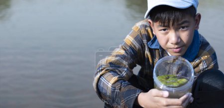 Foto de Asian boy holds transparent plastic tube which contains freshwater algae from river behind him inside, idea for learning water, river, environment, insect, fish, insecticide, pesticide and pollution. - Imagen libre de derechos