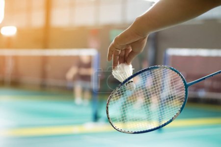 Photo pour Le joueur de badminton tient une raquette et un volant blanc crème devant le filet avant de le servir sur un autre côté du terrain. - image libre de droit