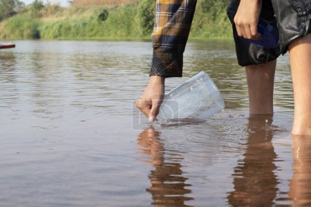 Téléchargez les photos : Écolier asiatique tenant un tube en plastique transparent pour garder l'eau douce de la rivière locale à l'intérieur pour étudier la fertilité de l'eau, de la rivière et des poissons et pour faire du travail de projet d'école de l'environnement en dehors des laboratoires. - en image libre de droit