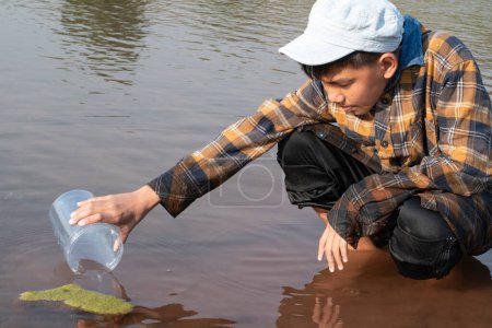 Téléchargez les photos : Écolier asiatique tenant un tube en plastique transparent pour garder l'eau douce de la rivière locale à l'intérieur pour étudier la fertilité de l'eau, de la rivière et des poissons et pour faire du travail de projet d'école de l'environnement en dehors des laboratoires. - en image libre de droit