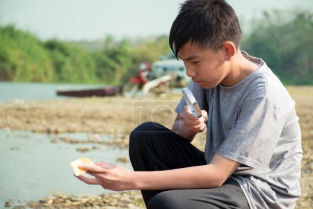 Foto de Asiático niño se sienta sosteniendo una lupa y mirando una lente convexa para tratar de quemar un pequeño pedazo de papel de los rayos del sol cerca de un río por la tarde durante las vacaciones escolares para hacer experimento. - Imagen libre de derechos