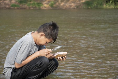 Foto de Niño asiático se sienta sosteniendo una lupa y mirando una lente de zoom para ver pequeñas criaturas submarinas en algear junto a un río por la tarde durante las vacaciones escolares para hacer experimentos. - Imagen libre de derechos