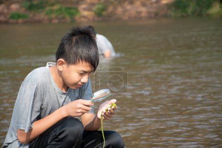 Foto de Niño asiático se sienta sosteniendo una lupa y mirando una lente de zoom para ver pequeñas criaturas submarinas en algear junto a un río por la tarde durante las vacaciones escolares para hacer experimentos. - Imagen libre de derechos