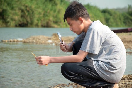 Foto de Asiático niño se sienta sosteniendo una lupa y mirando una lente convexa para tratar de quemar un pequeño pedazo de papel de los rayos del sol cerca de un río por la tarde durante las vacaciones escolares para hacer experimento. - Imagen libre de derechos