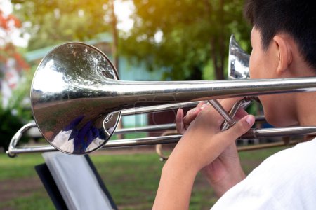 Téléchargez les photos : Instrument de trompette tenu dans les mains d'un étudiant asiatique qui s'entraîne avant de le jouer lors d'une cérémonie en l'honneur du drapeau national le matin. Focus doux et sélectif sur cloche circulaire de trompette. - en image libre de droit