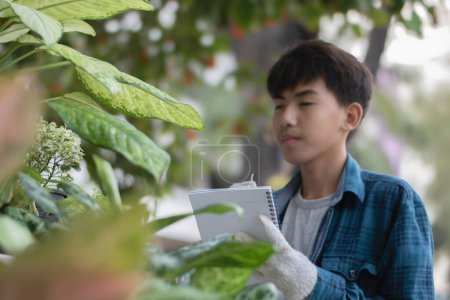 Asiático lindo chico sostiene pluma y portátil cerca de varias plantas de interior macetas y la grabación y el aprendizaje del cultivo de plantas de interior, enfoque suave y selectivo.