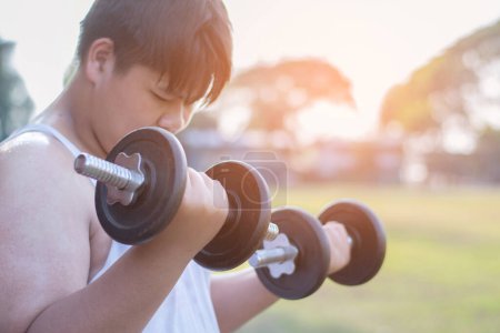 Young asian chubby boy doing exercise with dumbbells in outdoor park in the sunset time of the day, sunlight edited.