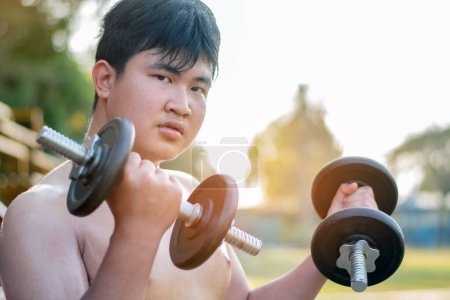 Young asian chubby boy doing exercise with dumbbells in outdoor park in the sunset time of the day, sunlight edited.