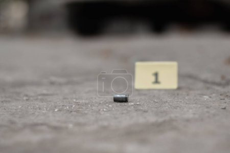 Pistolenkugelhülse auf Zementboden vor Nummer eins gelbem Papier in der Nähe von Autorädern, Konzept für Ermittlungen und Verbrechen unter Verwendung von Pistole, weicher Fokus.
