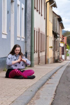 Téléchargez les photos : Lohnsfeld, Allemagne - Le 16 mai 2021 : Une adolescente assise les jambes croisées sur un trottoir de la rue principale à Lohnsfeld, Allemagne. - en image libre de droit