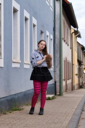 Téléchargez les photos : Lohnsfeld, Allemagne - Le 16 mai 2021 : Adolescente portant des collants roses et noirs debout sur un trottoir devant un bâtiment bleu à Lohnsfeld, Allemagne. - en image libre de droit