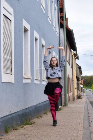 Téléchargez les photos : Lohnsfeld, Allemagne - Le 16 mai 2021 : Une adolescente équilibre sur une jambe avec les bras au-dessus de la tête debout sur un trottoir devant un bâtiment bleu un jour de printemps à Lohnsfeld, Allemagne. - en image libre de droit