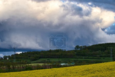 Photo for Light clouds and storm clouds over hill with trees next to a rapeseed field on a spring evening near Lohnsfeld, Germany. - Royalty Free Image