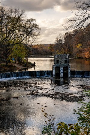 Photo for Morristown, NJ - USA - Nov 5, 2022 An autumnal vertical view of New Jersey's historic stone Speedwell Dam during sunset. Fly fisherman in the foreground and people hiking in the distance. - Royalty Free Image