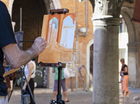 Nahaufnahme der Hand eines Malers, während er mit Pinsel auf Leinwand eine Outdoor-Szene auf einem kleinen Platz in Venedig, Italien, malt