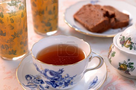 Photo for Close-up on a porcelain cup filled with tea. Slices of chocolate pound cake, two glasses filled with orange juice and a porcelain sugar bowl. All items placed on a floral tablecloth. - Royalty Free Image