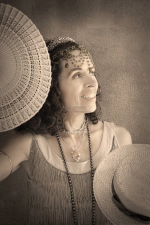Photo for Portrait of a smiling woman dressed in 1920s style with fringe flapper dress and jewelry. She is holding a fan and a straw boater hat. Vintage sepia old photo effect. - Royalty Free Image