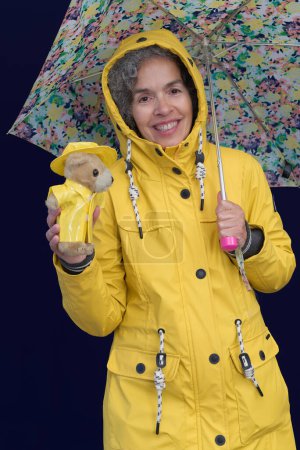 Téléchargez les photos : Femme aux cheveux gris est préparé pour les jours de pluie. Elle et son ours en peluche portent des imperméables jaunes. Elle tient un parapluie ouvert. Fond sombre. - en image libre de droit