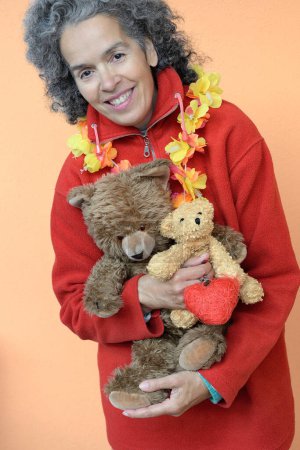 Photo for A woman over 50 with curly graying hair is holding two Teddy bears in her arms and a red heart in one hand. Light shade of orange background. - Royalty Free Image